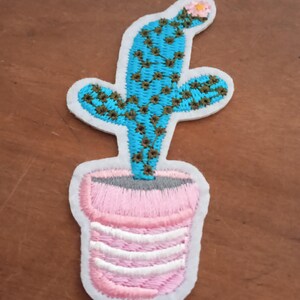 Peut inclure: Écusson brodé représentant un cactus turquoise avec des détails marron et une petite fleur rose, dans un pot rose et blanc rayé. Le patch a une bordure blanche, idéal pour les vêtements ou accessoires.