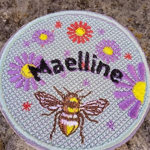 Peut inclure: Écusson brodé avec un fond bleu clair, représentant une abeille et le nom "Maelline" en noir. Des fleurs violettes et jaunes et des étoiles rouges ornent le nom et l'abeille. L'écusson a une bordure violet clair.