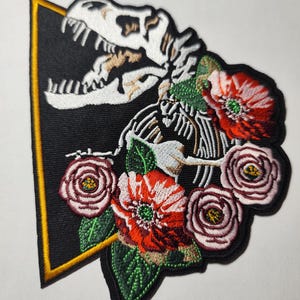 Peut inclure: Un écusson brodé représentant un squelette de dinosaure T-Rex blanc sur un fond triangulaire noir avec une bordure jaune. Le squelette est entouré de fleurs rouges et roses détaillées avec des feuilles vertes, formant un arrangement floral.