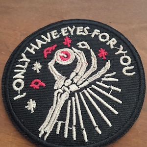 Peut inclure: Écusson rond noir avec l'inscription blanche "I ONLY HAVE EYES FOR YOU". Il représente une main squelettique tenant un œil, avec des détails rouges et blancs.