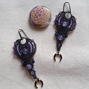 Peut inclure: Boucles d'oreilles macramé faites à la main, ornées de perles d'améthyste et d'un croissant de lune. Les boucles d'oreilles sont noires et violettes, avec un motif en forme de cœur. Une pierre violette avec un motif doré est également présente.