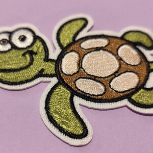 Peut inclure: Écusson brodé en forme de tortue souriante, avec une tête et des membres verts, et une carapace marron et beige. Le patch a une bordure blanche, pour vêtements ou accessoires.