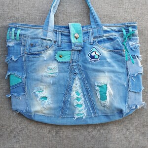 以下が含まれることがあります： 「ワカナ」と書かれたパッチが付いた、ターコイズ色のトリムが付いた青いデニムのトートバッグ。バッグは、いくつかの裂け目や破れがある、使い古した外観をしています。