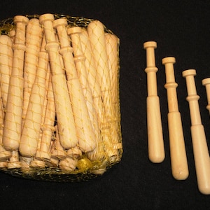May include: A collection of light-colored wooden bobbins, some inside a yellow mesh bag, and others arranged separately. The bobbins have a distinctive shape with a rounded base and a cylindrical body, ideal for lace making.
