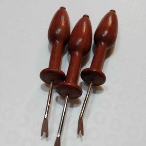 May include: Three wooden tools with a dark brown finish. Each tool has a rounded top, a circular base, and a metal shaft ending in a forked tip. The tools are arranged on a white surface.