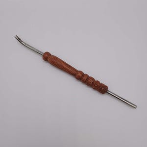 Puede incluir: Una herramienta de madera y metal con un extremo curvado y un mango decorativo. El mango es de madera marrón oscuro con una serie de crestas talladas. La parte metálica es plateada y se extiende desde ambos extremos.