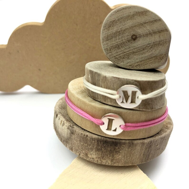 Puede incluir: Dos pulseras personalizadas con las letras "M" y "L" en dijes de plata. Las pulseras est&aacute;n en rondas de madera con un cord&oacute;n rosa y blanco.