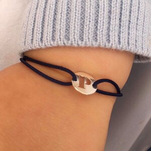 Puede incluir: Una pulsera el&aacute;stica negra con un dije plateado con la letra "P" en un dise&ntilde;o recortado.