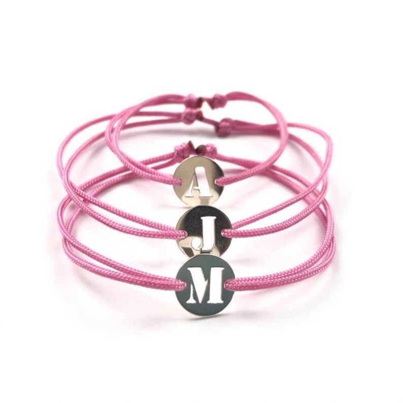 Puede incluir: Tres pulseras de amistad de color rosa con dijes plateados con las letras A, J y M.