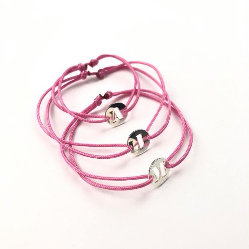 Puede incluir: Tres pulseras de amistad de color rosa con dijes plateados con las letras A, J y M.