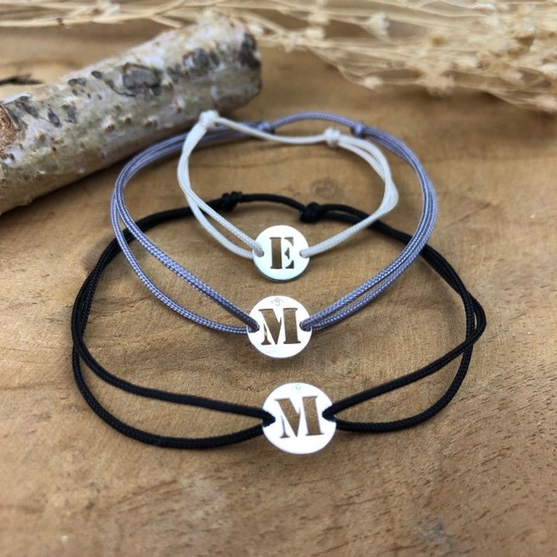 Puede incluir: Tres pulseras ajustables con dijes de plata con las letras E y M. Las pulseras est&aacute;n hechas de cord&oacute;n negro, gris y blanco.