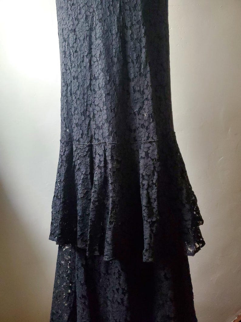 May include: A floor-length black lace dress with a tiered skirt. The dress has a fitted bodice and a flared skirt, creating an elegant silhouette. The intricate lace pattern adds texture and visual interest to the garment.