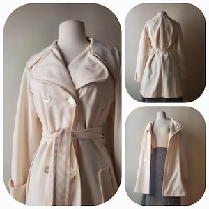 Puede incluir: Gabardina color crema con diseño de doble botonadura, con cuello con solapas, cierres de botones y cinturón a juego. El abrigo se exhibe en un maniquí, mostrando su ajuste a medida y su estilo clásico. El abrigo mide aproximadamente 91 cm de largo.