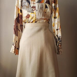 Puede incluir: Un conjunto vintage de dos piezas con una blusa de manga larga y una falda. La blusa tiene un fondo crema con un estampado abstracto colorido. La falda es de color crema liso, de cintura alta y silueta acampanada.