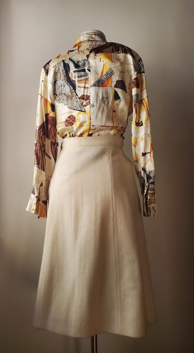 Puede incluir: Un conjunto vintage de dos piezas con una blusa de manga larga y una falda. La blusa tiene un fondo crema con un estampado abstracto colorido. La falda es de color crema liso, de cintura alta y silueta acampanada.