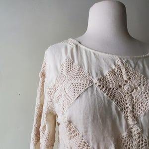 May include: Cream-colored long-sleeved top with crochet detailing. The top features a round neckline and is adorned with diamond-shaped crochet patches in a lighter cream hue. The sleeves also have crochet accents, creating a textured, bohemian look.