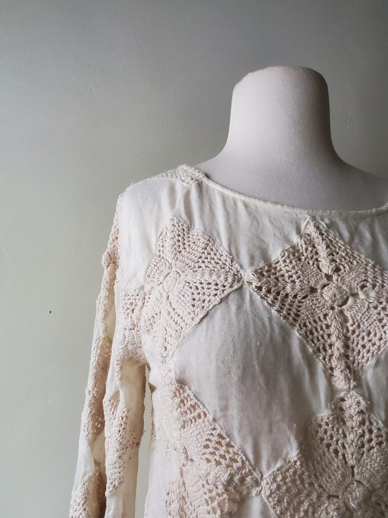 May include: Cream-colored long-sleeved top with crochet detailing. The top features a round neckline and is adorned with diamond-shaped crochet patches in a lighter cream hue. The sleeves also have crochet accents, creating a textured, bohemian look.