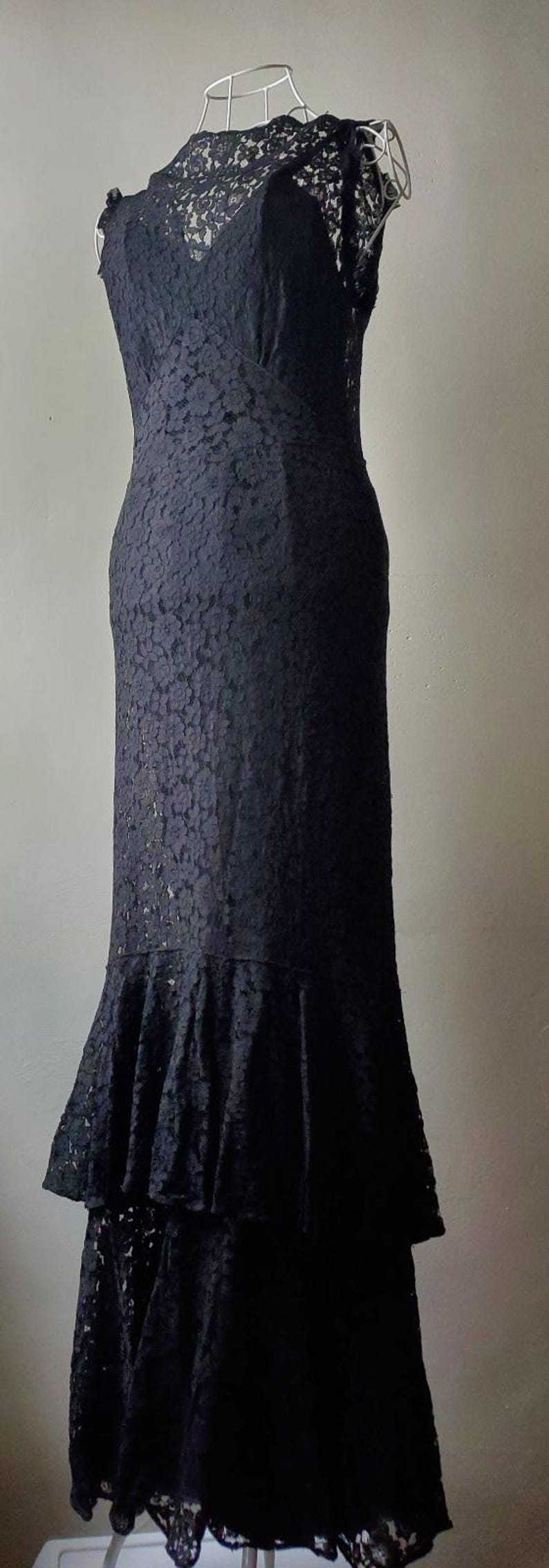 May include: A black lace dress displayed on a mannequin. The dress features a fitted bodice, a flared skirt with a ruffled hem, and a delicate lace overlay. The dress is floor-length and suitable for formal occasions.