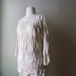 May include: Off-white crochet top with long sleeves and a round neckline. The top features a detailed floral crochet pattern and a scalloped hemline. The garment is displayed on a mannequin.