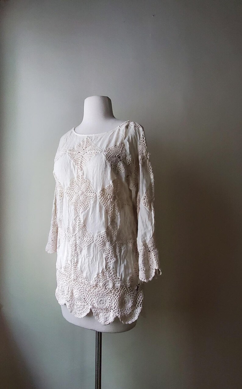 May include: Off-white crochet top with long sleeves and a round neckline. The top features a detailed floral crochet pattern and a scalloped hemline. The garment is displayed on a mannequin.