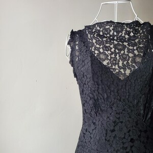 May include: A black lace dress displayed on a white dress form. The dress features a V-neckline and a floral lace pattern. The dress is sleeveless with a fitted bodice and a flared skirt.