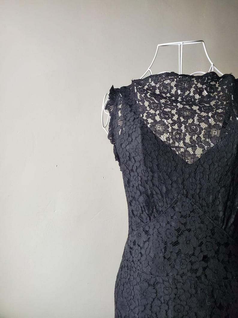 May include: A black lace dress displayed on a white dress form. The dress features a V-neckline and a floral lace pattern. The dress is sleeveless with a fitted bodice and a flared skirt.