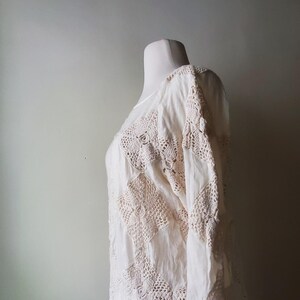 May include: Cream-colored, long-sleeved top with intricate crochet detailing. The garment features a patchwork design with square crochet panels and a flowing, lightweight fabric. The top is displayed on a mannequin, showcasing its texture and design.