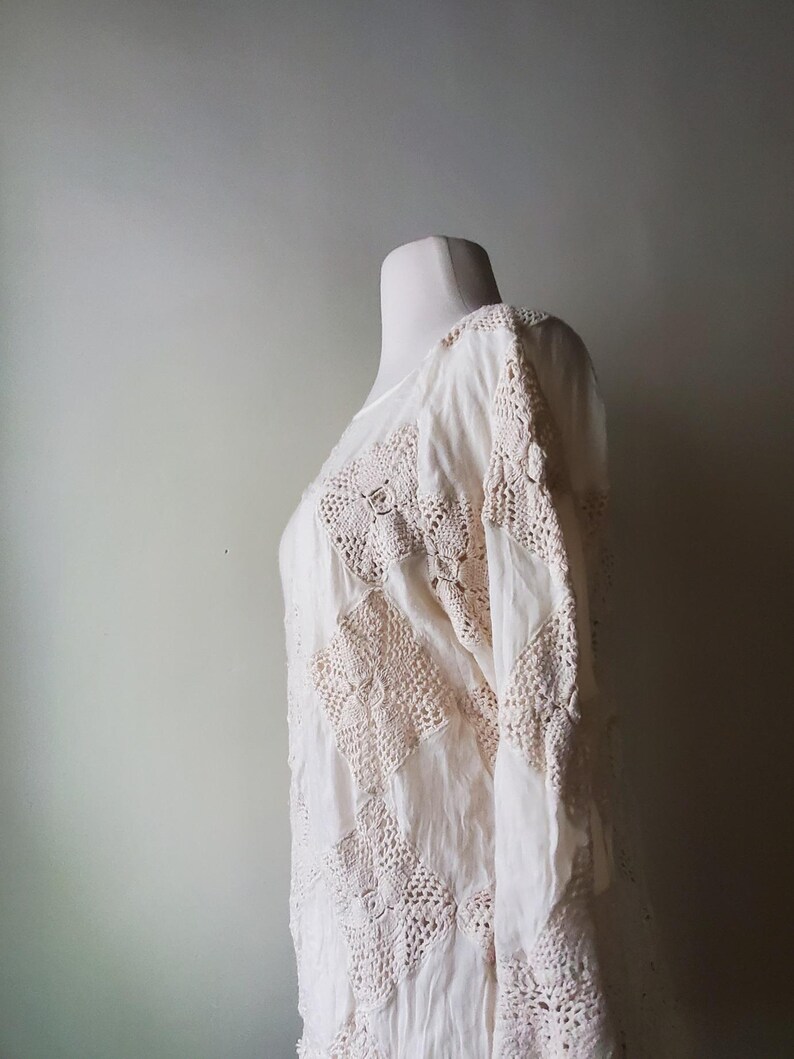 May include: Cream-colored, long-sleeved top with intricate crochet detailing. The garment features a patchwork design with square crochet panels and a flowing, lightweight fabric. The top is displayed on a mannequin, showcasing its texture and design.