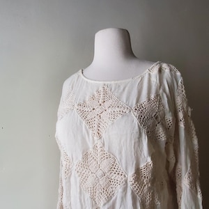 May include: Cream-colored, long-sleeved top with a round neckline. The top features a patchwork design of crocheted squares and panels, creating a textured, vintage aesthetic. The fabric appears lightweight and airy, suitable for warmer weather.