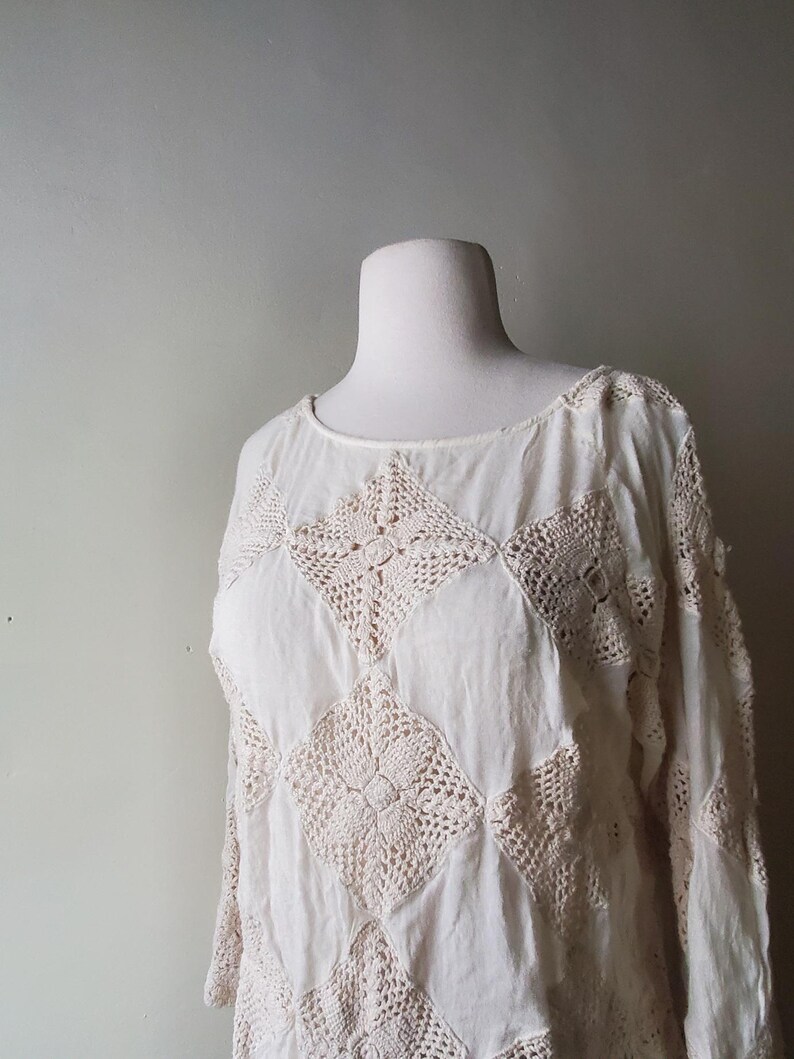 May include: Cream-colored, long-sleeved top with a round neckline. The top features a patchwork design of crocheted squares and panels, creating a textured, vintage aesthetic. The fabric appears lightweight and airy, suitable for warmer weather.
