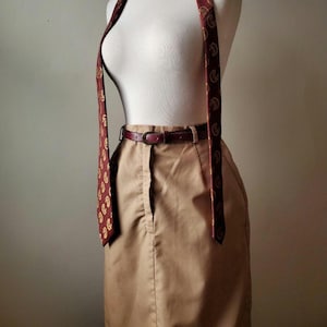 May include: A tan skirt with a burgundy leather belt and a burgundy paisley tie draped over a mannequin. The tie has a gold paisley pattern. The skirt is knee-length and has a button and zip closure.