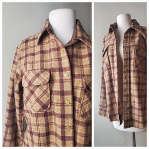 Pode incluir: Uma camisa vintage abotoada com um padrão xadrez bege, bordô e creme. A camisa tem dois bolsos no peito e uma gola. A camisa de manga comprida é exibida em um manequim.