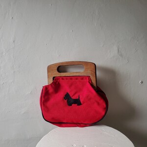 May include: A red handbag with a wooden handle and a black Scottie dog applique. The bag has a rounded shape and a black trim. The handle is a light brown color. The bag is sitting on a white surface.