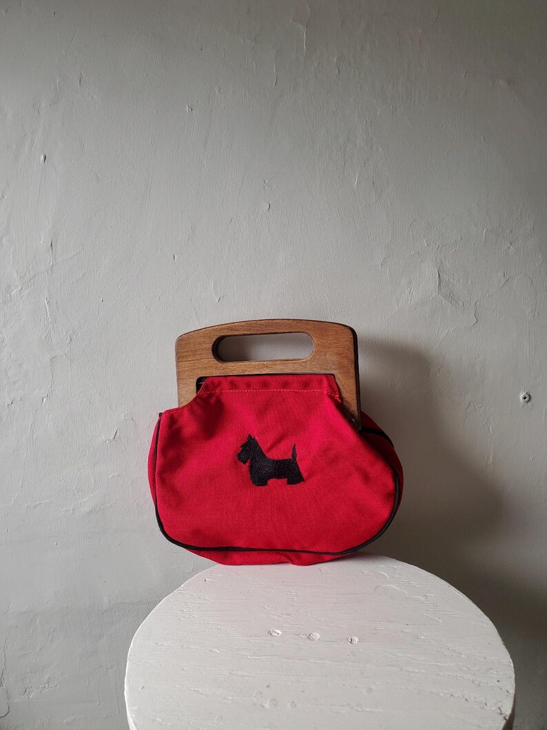 May include: A red handbag with a wooden handle and a black Scottie dog applique. The bag has a rounded shape and a black trim. The handle is a light brown color. The bag is sitting on a white surface.