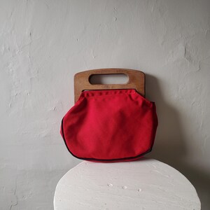 May include: A vintage red handbag with a wooden handle. The bag is a vibrant red color with a black trim. The wooden handle has a rectangular opening for carrying. The bag is sitting on a white, round table.