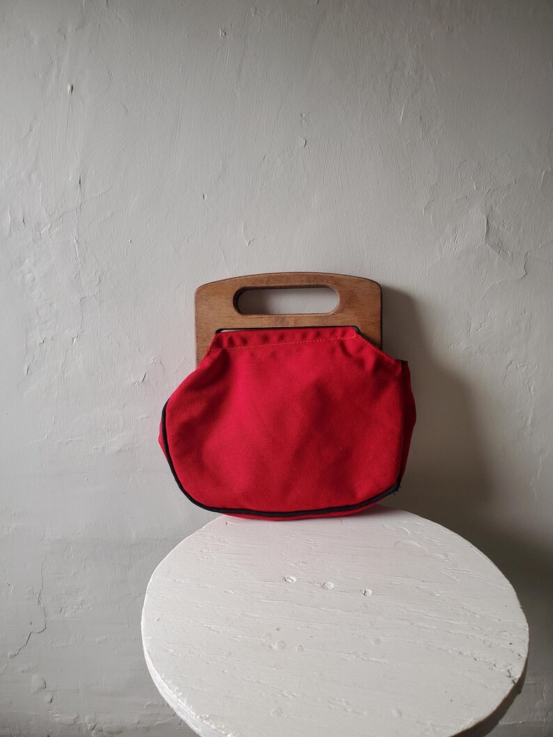 May include: A vintage red handbag with a wooden handle. The bag is a vibrant red color with a black trim. The wooden handle has a rectangular opening for carrying. The bag is sitting on a white, round table.