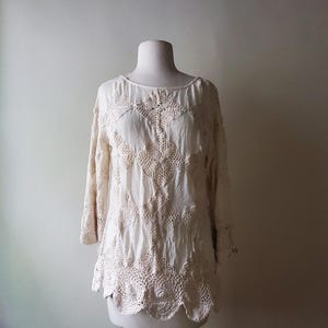 May include: Cream-colored, long-sleeved top with intricate crochet detailing. The blouse features a round neckline and a scalloped hem. The crochet design is visible across the bodice and sleeves, creating a textured, bohemian look.