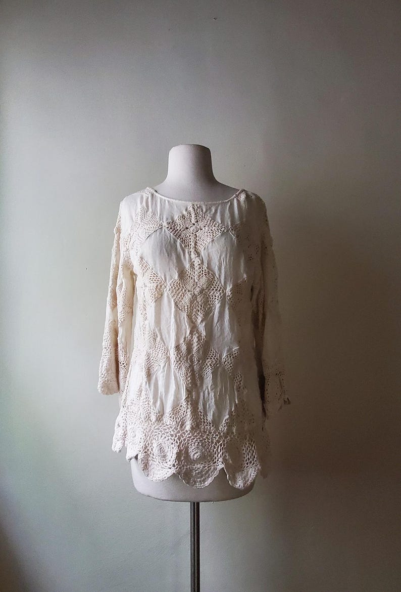 May include: Cream-colored, long-sleeved top with intricate crochet detailing. The blouse features a round neckline and a scalloped hem. The crochet design is visible across the bodice and sleeves, creating a textured, bohemian look.