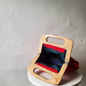 May include: A vintage handbag with a wooden frame and a red fabric exterior. The interior features a green and blue plaid pattern. The bag is open, revealing the plaid lining. The wooden frame has two handle openings.