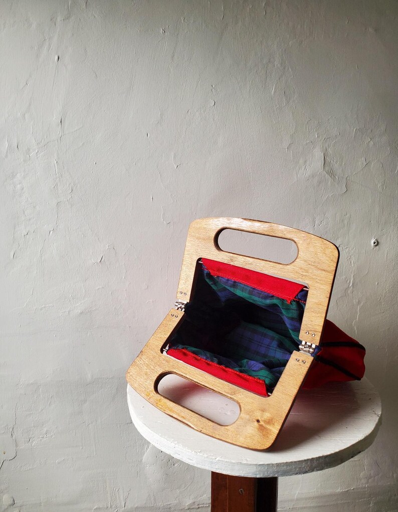 May include: A vintage handbag with a wooden frame and a red fabric exterior. The interior features a green and blue plaid pattern. The bag is open, revealing the plaid lining. The wooden frame has two handle openings.
