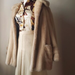 Puede incluir: Un conjunto vintage con un abrigo de piel sint&eacute;tica color crema con solapas grandes y bolsillos, sobre una blusa estampada y una falda a juego. La blusa tiene un estampado ecuestre marr&oacute;n y crema. La falda es de color crema, con dise&ntilde;o plisado.