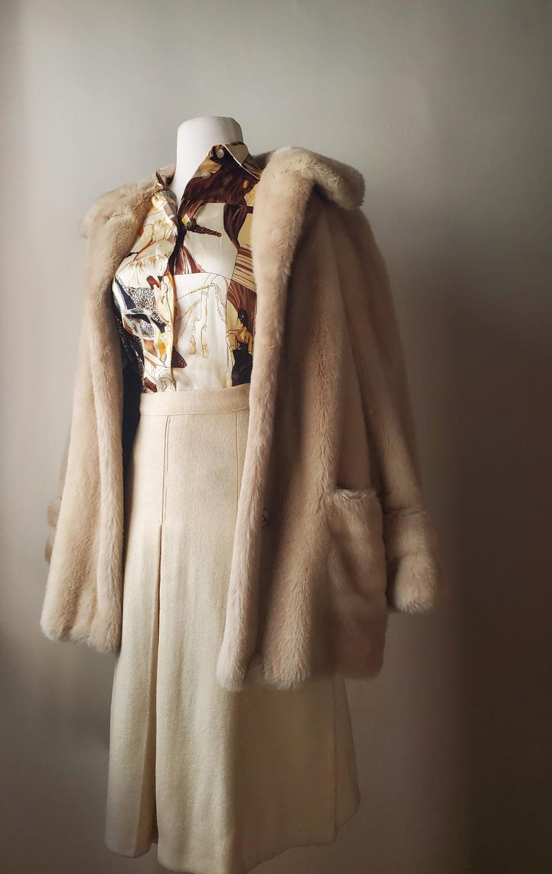 Puede incluir: Un conjunto vintage con un abrigo de piel sint&eacute;tica color crema con solapas grandes y bolsillos, sobre una blusa estampada y una falda a juego. La blusa tiene un estampado ecuestre marr&oacute;n y crema. La falda es de color crema, con dise&ntilde;o plisado.