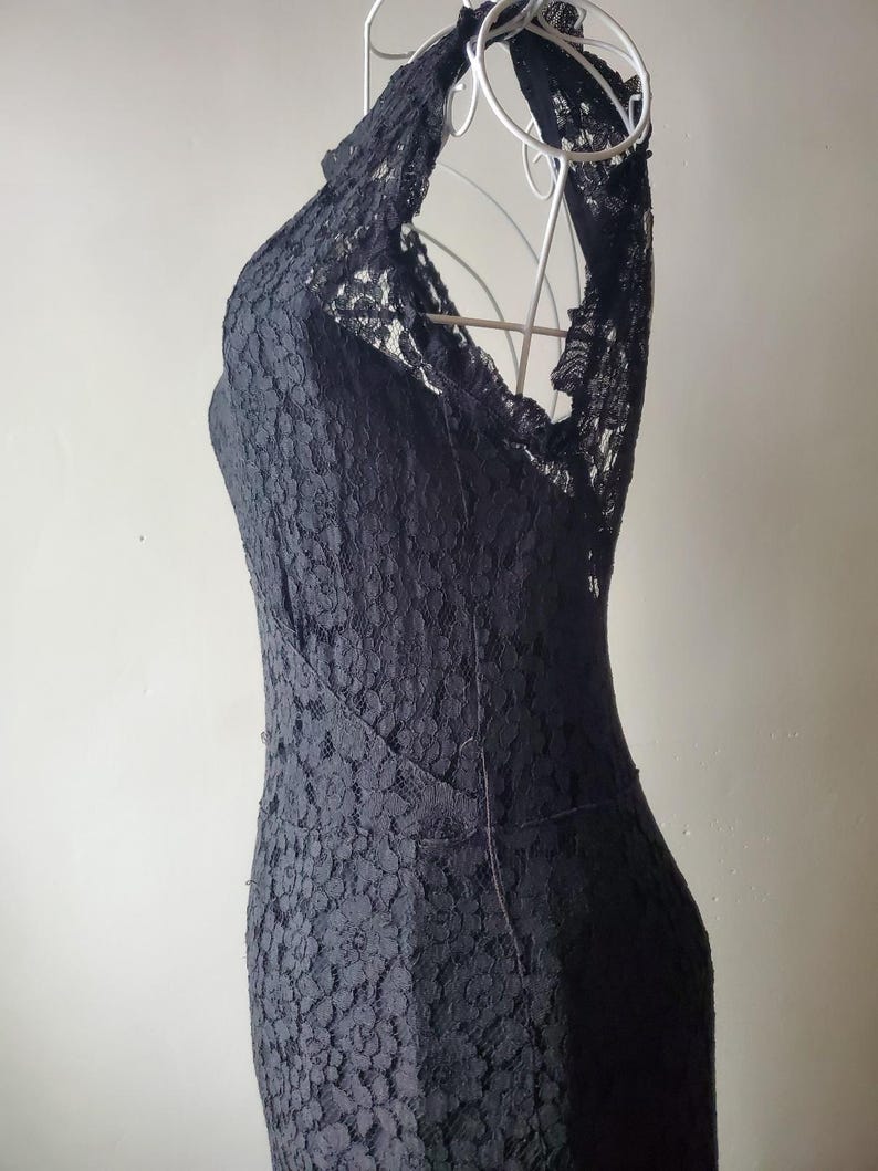 May include: A black lace dress with a halter neckline. The dress is displayed on a white wire dress form. The dress has a fitted silhouette and a floral lace pattern. The dress is a formal wear item.