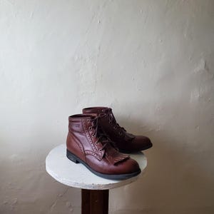 May include: A pair of brown leather ankle boots with laces and fringe detail. The boots are displayed on a small, round, white table. The boots have a classic, vintage style.