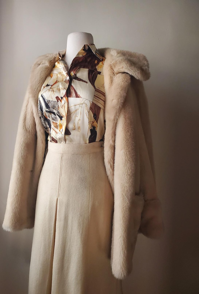 Puede incluir: Un abrigo de piel sint&eacute;tica color crema, una camisa con botones estampada y una falda a juego se exhiben en un maniqu&iacute;. La camisa presenta un estampado con elementos marrones, beige y negros. La falda tiene una abertura frontal y el abrigo tiene un cuello ancho.
