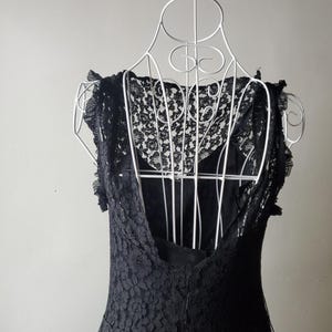 May include: A black lace dress displayed on a white wire dress form. The dress features a low back and delicate lace detailing on the bodice and sleeves. The dress is sleeveless and appears to be made of a soft, flowing fabric.
