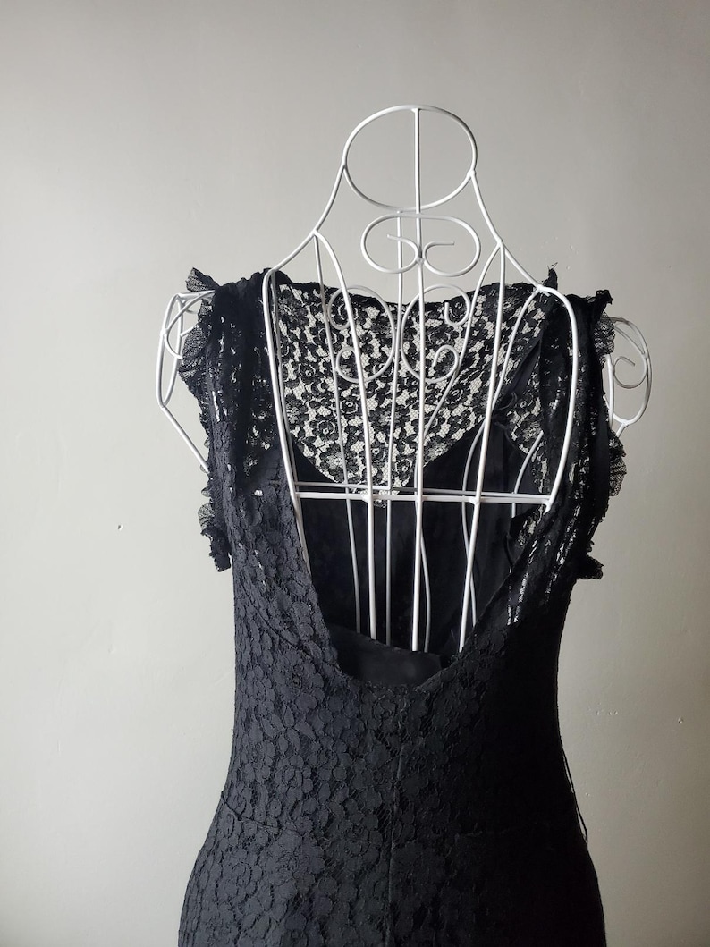 May include: A black lace dress displayed on a white wire dress form. The dress features a low back and delicate lace detailing on the bodice and sleeves. The dress is sleeveless and appears to be made of a soft, flowing fabric.