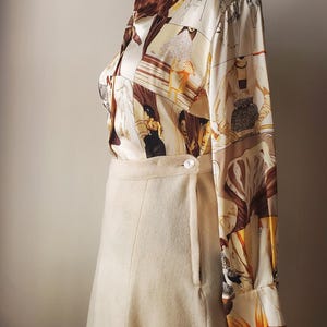 Puede incluir: Un conjunto vintage con una falda color crema y una blusa estampada. La blusa de manga larga muestra un dise&ntilde;o repetido con figuras en tonos marr&oacute;n, beige y naranja. La falda tiene cierre de bot&oacute;n y una forma sutil de l&iacute;nea A.