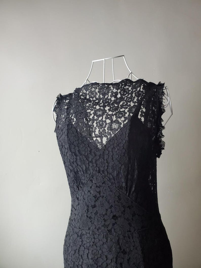 May include: A black lace dress displayed on a wire dress form. The dress features a high neckline and a fitted bodice, with a flared skirt. The intricate floral lace pattern covers the entire dress, creating a delicate and elegant look.
