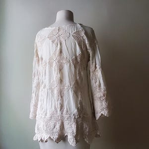 May include: A white, long-sleeved top with intricate crochet detailing. The top features a round neckline, bell sleeves, and a scalloped hem. The crochet design is a lighter shade of white, creating a subtle contrast. The garment is displayed on a mannequin.
